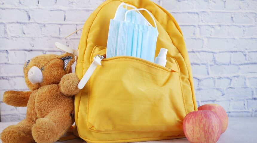 A bright yellow daycare bag packed with essentials including face masks, sanitizer spray, a soft teddy bear, and apples—perfect examples of daycare bag essentials for young children.