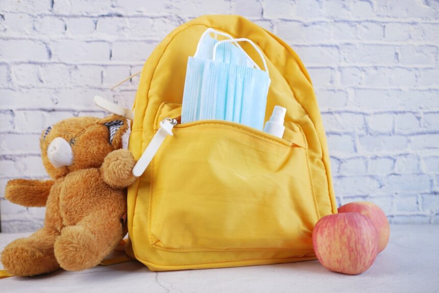 A bright yellow daycare bag packed with essentials including face masks, sanitizer spray, a soft teddy bear, and apples—perfect examples of daycare bag essentials for young children.